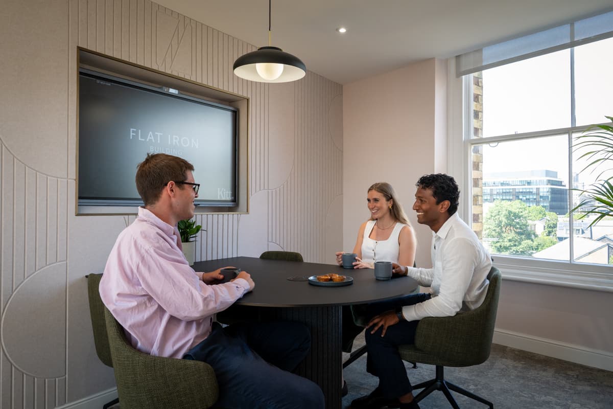 Three colleagues around a meeting table in a Kitt workspace, with daylight from a tall window behind them.